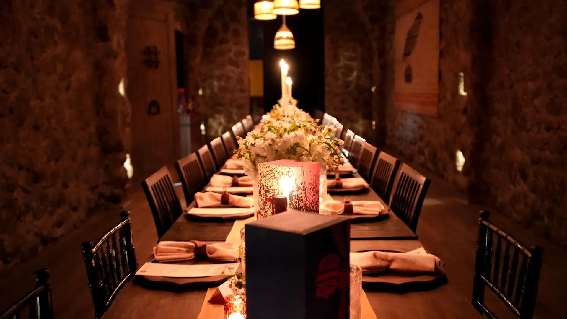 Long dining table set for an immersive tasting experience, featuring elegant floral arrangements and ambient lighting in a rustic stone setting.